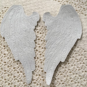 May include: A pair of white fabric angel wings with a decorative, embroidered design. The wings are on a white crocheted surface.