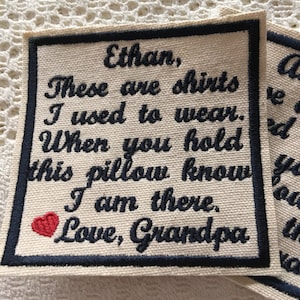Op de afbeelding: Een wit vierkant van stof met zwarte geborduurde tekst die luidt: "Ethan, Dit zijn shirts die ik droeg. Wanneer je dit kussen vasthoudt, weet dan dat ik er ben. Liefs, Opa". Een rood hart is geborduurd in de linkerbovenhoek.