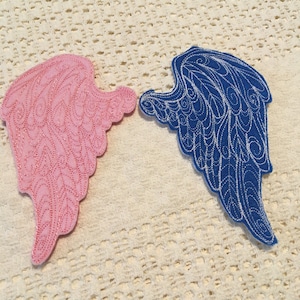 May include: Two felt angel wings, one pink and one blue, with intricate embroidered designs. The wings are laid out on a white crocheted surface.