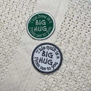 Op de afbeelding: Twee ronde geborduurde patches met de tekst "This bear is a big hug from me to you" en "This quilt is a big hug from me to you". De eerste patch is groen met witte tekst en de tweede patch is wit met zwarte tekst.