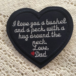 May include: A navy blue heart-shaped patch with white embroidery that reads "I love you a bushel and a peck, with a hug around the neck. Love, Dad". A small red heart is embroidered next to the word "Love".