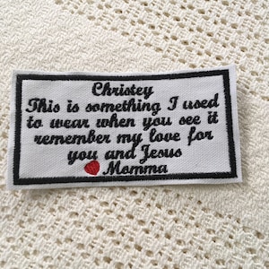 Może przedstawiać: Biały materiałowy znaczek z czarnym haftem, na którym widnieje napis "Christey This is something I used to wear when you see it remember my love for you and Jesus Momma" z czerwonym sercem.