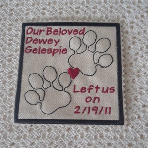 Puede incluir: Un parche conmemorativo de tela blanca con costuras negras. Presenta dos huellas de patas negras con un corazón rojo en el centro. El texto "Our Beloved Dewey Gelespie Left us on 2/19/11" está bordado en rojo.