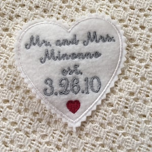 May include: White heart-shaped fabric patch with silver embroidery that reads "Mr. and Mrs. Minonna est. 3.26.10". A small red heart is embroidered below the date.