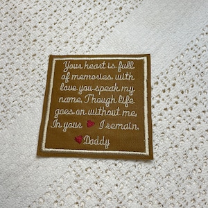 Może przedstawiać: Brązowy materiałowy znaczek z białym haftem, na którym widnieje tekst: "Your heart is full of memories, with love you speak my name. Though life goes on without me, In your I remain, Daddy". Są dwa czerwone haftowane serca.
