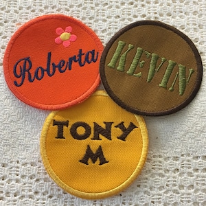Pode incluir: Três patches redondos de tecido com nomes bordados. Os patches são laranja, castanho e amarelo. Os nomes são Roberta, Kevin e Tony M.