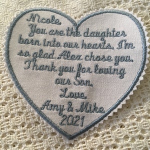 May include: A white heart-shaped fabric patch with blue embroidered text that reads "Nicole You are the daughter born into our hearts. I'm so glad Alex chose you. Thank you for loving our Son Love Amy & Mike 2021".