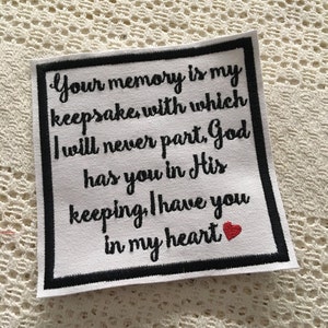 Op de afbeelding: Witte vierkante stof met een zwarte rand en zwarte geborduurde tekst die luidt "Your memory is my keepsake with which I will never part, God has you in His keeping, I have you in my heart". Een klein rood hart is aan het einde van de tekst geborduurd.