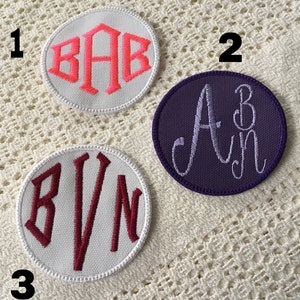 Peut inclure: Trois cercles en tissu blanc avec des lettres brodées. Le premier cercle a les lettres "BAB" en fil rose. Le deuxième cercle a les lettres "AB" en fil violet. Le troisième cercle a les lettres "BVN" en fil marron.