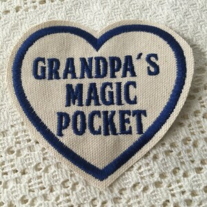Puede incluir: Un parche blanco en forma de corazón con un borde bordado azul. El parche dice "Grandpa's Magic Pocket" en bordado azul.