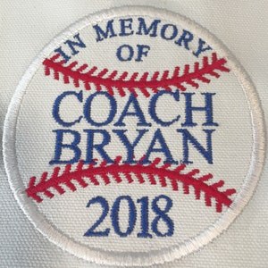 May include: A white baseball patch with red stitching and blue text. The text reads "IN MEMORY OF COACH BRYAN 2018". The patch is round and has a stitched border, resembling a baseball.