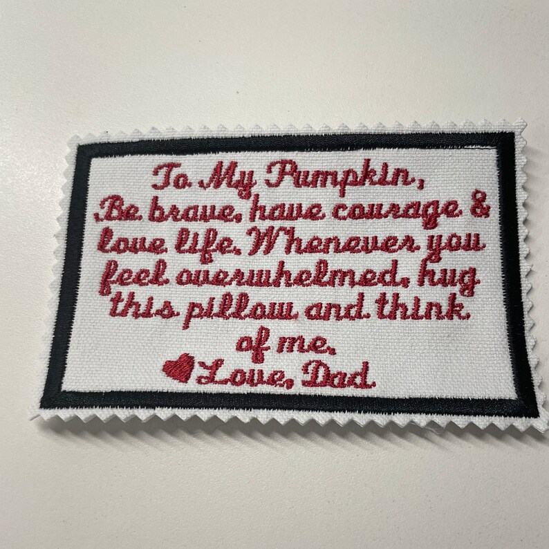 Op de afbeelding: Een wit stoffen kussen met rode geborduurde tekst die luidt "To My Pumpkin, Be brave, have courage & love life. Whenever you feel overwhelmed, hug this pillow and think of me. Love, Dad".