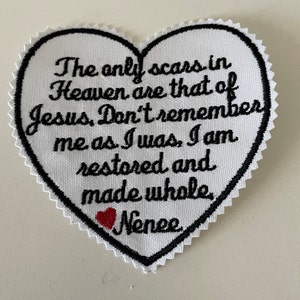 Puede incluir: Parche de tela blanco en forma de corazón con texto bordado en negro que dice "The only scars in Heaven are that of Jesus, Don't remember me as I was, I am restored and made whole." Un pequeño corazón rojo está bordado debajo del texto con el nombre "Nenee" bordado debajo del corazón.