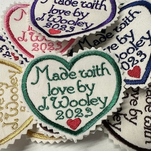 May include: A close-up of several heart-shaped fabric patches with green, blue, purple, and pink embroidery. The patches say "Made with love by J. Wooley 2023".