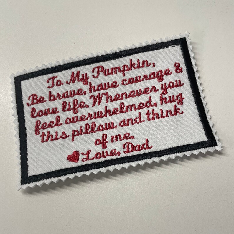 Op de afbeelding: Witte stoffen patch met een zwarte rand en rode geborduurde tekst. De tekst luidt "To My Pumpkin, Be brave, have courage & love life. Whenever you feel overwhelmed, hug this pillow and think of me. Love, Dad".