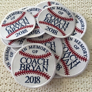 Könnte beinhalten: Ein Stapel weißer, baseballförmiger Patches mit roten Nähten und blauem Text. Der Text lautet "In Memory of Coach Bryan 2018".