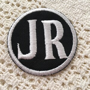 Pu&ograve; includere: Una toppa ricamata in bianco e nero con le lettere "JR" in un cerchio.