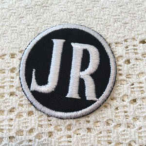 Pu&ograve; includere: Una toppa ricamata in bianco e nero con le iniziali "JR" in un cerchio bianco su sfondo nero.