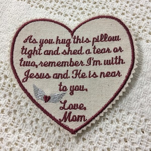 Può includere: Un cerotto ricamato a forma di cuore con un messaggio di conforto e amore. Il testo recita: "As you hug this pillow tight and shed a tear or two, remember I'm with Jesus and He is near to you. Love, Mom".