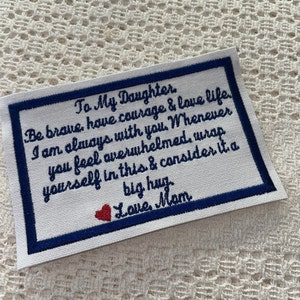 Może przedstawiać: Biały materiał z niebieską obwódką i czerwonym sercem. Materiał ma wyszyte przesłanie, które brzmi: "To My Daughter Be brave, have courage & love life. I am always with you. Whenever you feel overwhelmed, wrap yourself in this & consider it a big hug Love Mom"