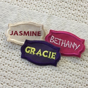 Könnte beinhalten: Drei bestickte Stoff-Namensschilder mit dekorativen Rändern. Die Schilder sind beige, rosa und lila. Die Namen auf den Schildern sind "Jasmine", "Bethany" und "Gracie".