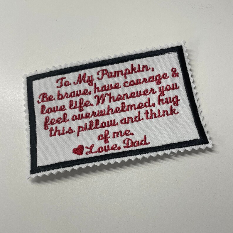 Op de afbeelding: Witte stoffen patch met zwarte stiksels en een rood geborduurd bericht dat luidt "To My Pumpkin, Be brave, have courage & love life. Whenever you feel overwhelmed, hug this pillow and think of me. Love, Dad".
