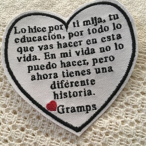 May include: A white heart-shaped patch with black stitching and a red heart in the bottom right corner. The patch has a message in black text that reads: "Lo hice por ti mija, tu educación, por todo lo que vas hacer en esta vida. En mi vida no lo puedo hacer, pero ahora tienes una diferente historia. Gramps"