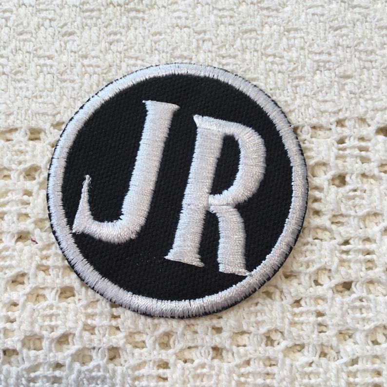 Pu&ograve; includere: Una toppa ricamata in bianco e nero con le lettere "JR" in un cerchio bianco su sfondo nero.