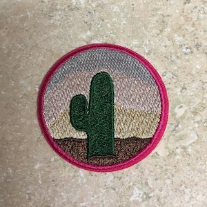 Op de afbeelding: Een ronde geborduurde patch met een roze rand. De patch toont een groene cactus met een bruin woestijnlandschap en een meerkleurige zonsondergang op de achtergrond.