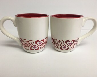 以下が含まれることがあります： 赤い縁取りと底にピンクと赤のハート模様がついた白いセラミック製のマグカップ2つ
