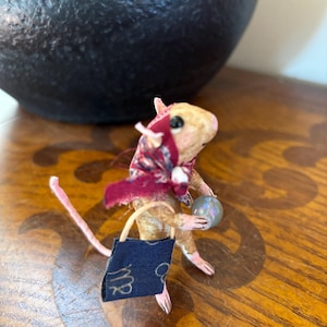 May include: A small tan mouse figurine wearing a dark red patterned headscarf. The mouse holds a small iridescent sphere in one hand and carries a dark blue rectangular bag with the gold zodiac symbol for Virgo printed on the front.