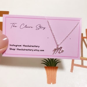 Puede incluir: Un collar de plata con un colgante de la letra "M" en cursiva. El collar se muestra en una tarjeta rosa con el texto "The Claire Story" e información de redes sociales.