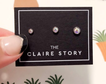 Puede incluir: Un conjunto de tres pendientes de botón iridiscentes sobre una tarjeta negra con el texto "THE CLAIRE STORY" en blanco. Los pendientes son de diferentes tamaños, con un botón pequeño, mediano y grande. El fondo es de color claro con una ilustración de planta.