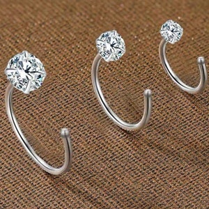 May include: Three silver nose rings with clear cubic zirconia stones. The rings are in different sizes and are arranged in a row.