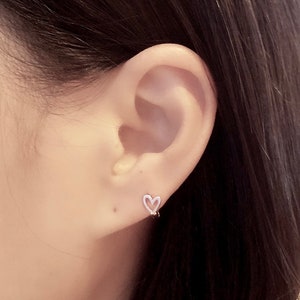 May include: A silver heart-shaped earring with a simple design. The earring is worn in the left ear of a person with long dark hair.