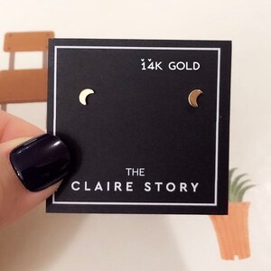 May include: A pair of gold crescent moon stud earrings on a black card. The card reads "14K GOLD" and "THE CLAIRE STORY" in white text. The earrings are small and delicate, with a polished finish.