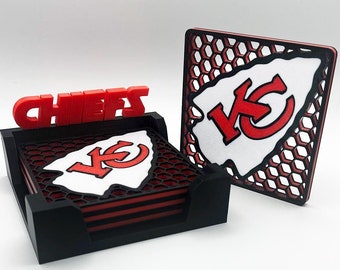 Könnte beinhalten: Ein Satz mit vier schwarzen Untersetzern mit einem rot-weißen Kansas City Chiefs Logo. Die Untersetzer befinden sich in einem schwarzen Halter mit dem Wort "CHEERS" in roten Buchstaben. Es gibt auch einen einzelnen Untersetzer mit dem gleichen Logo auf einem schwarz-roten Wabenhintergrund.