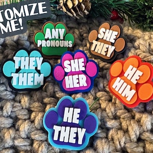 May include: A collection of colorful paw-shaped accessories, each displaying pronouns like "She/Her," "He/Him," and "They/Them." The accessories are in various colors, including blue, pink, orange, and brown, with white text. The phrase "Customize Me!" is also visible.