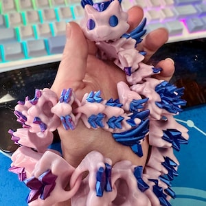 May include: A pink and blue 3D printed dragon bracelet with a crystal-like design. The bracelet is made up of multiple connected pieces, each with a unique shape and texture.