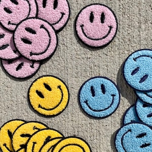 May include: A collection of colourful smiley face patches in yellow, pink and blue. The patches are made of fuzzy fabric and have a black outline.