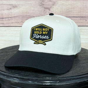 Puede incluir: Gorra de béisbol blanca y negra con el texto "I WILL NOT HOLD MY Horses" en un parche dorado y negro. La gorra tiene una visera negra y un botón negro. El sombrero está sobre una superficie oscura y redonda.