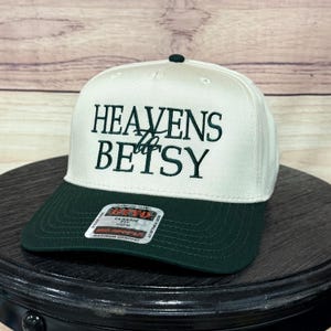 Op de afbeelding: Een witte en donkergroene baseballcap met de geborduurde tekst "HEAVENS Betsy". De cap heeft een gebogen klep en een knoop aan de bovenkant. Gemaakt van premium stof, biedt het een klassieke pasvorm.