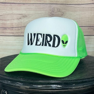 May include: A neon green and white trucker hat with the word "WEIRD" in black block letters. An embroidered green alien head is on the right side. The hat has a green brim and mesh back.