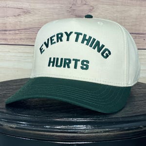 May include: A white and green baseball cap with the text "EVERYTHING HURTS" embroidered in green on the front.