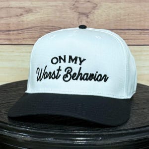 Può includere: Un cappellino da baseball bianco con visiera nera e bottone nero. La frase "ON MY Worst Behavior" è ricamata in nero sul davanti. Il cappello è esposto su una superficie scura, con sfondo in legno.