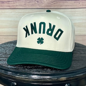 Puede incluir: Una gorra de béisbol blanca y verde con la palabra "DRUNK" bordada en verde en la parte delantera. Un trébol verde se encuentra debajo de la palabra.