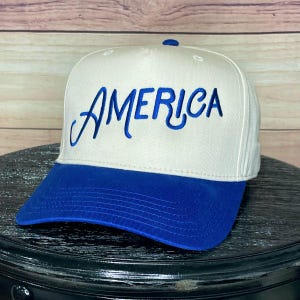Puede incluir: Gorra de béisbol blanca y azul con la palabra "AMERICA" bordada en azul en la parte delantera. La gorra tiene una visera azul y una corona blanca, con un pequeño botón azul en la parte superior.