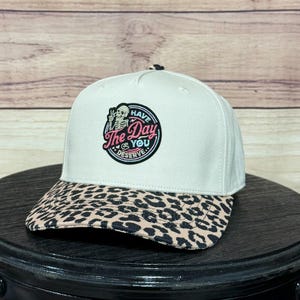 Puede incluir: Gorra de béisbol blanca con visera con estampado de leopardo. La gorra presenta un parche circular con un esqueleto haciendo la señal de la paz y el texto "Have The Day You Deserve".