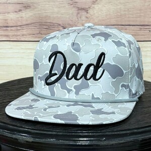 Könnte beinhalten: Grau-weißes Baseball-Cap mit Tarnmuster und dem Wort "Dad" in schwarzer Schrift auf der Vorderseite. Die Kappe hat einen flachen Schirm und eine graue Kordel vorne.