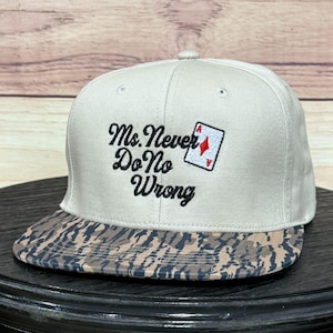 Puede incluir: Gorra de béisbol blanca con visera plana. Presenta el texto bordado "Ms. Never Do No Wrong" en escritura negra, junto con una carta de juego con un diamante rojo. La visera tiene un patrón de camuflaje.
